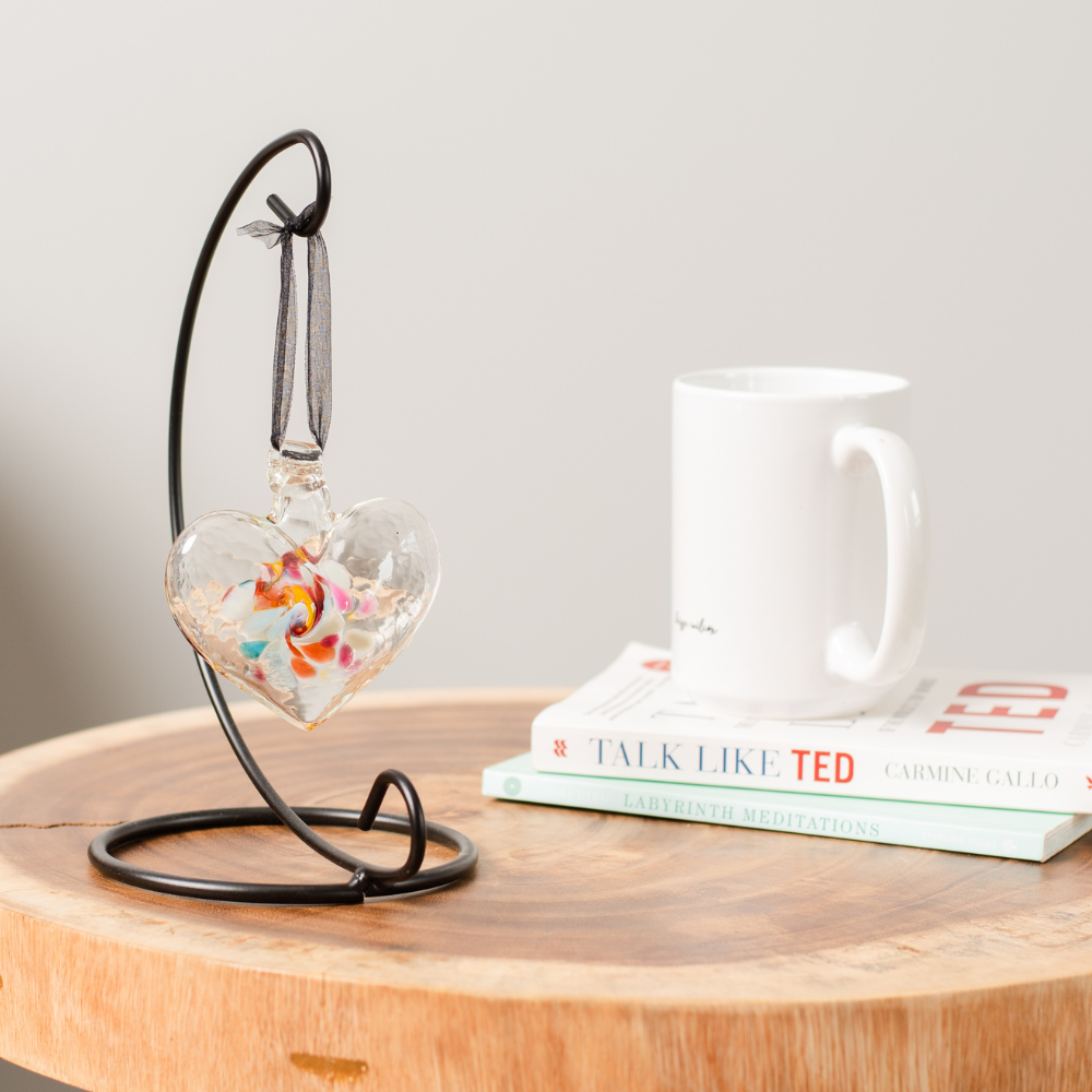 Glass heart with pink, blue, orange, yellow and opaque white swirled in the entre hanging from a black ribbon on a black curved single ornament holder. Displayed on a wood table with a white book and mug to the right. 