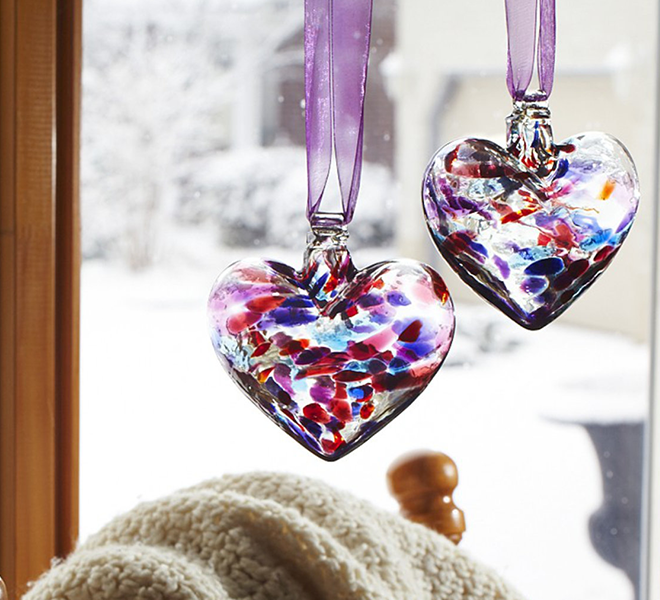 Two glass hearts with pink, purple and blue glass mosaiced across the glass hanging in front of a window from purple ribbon. 