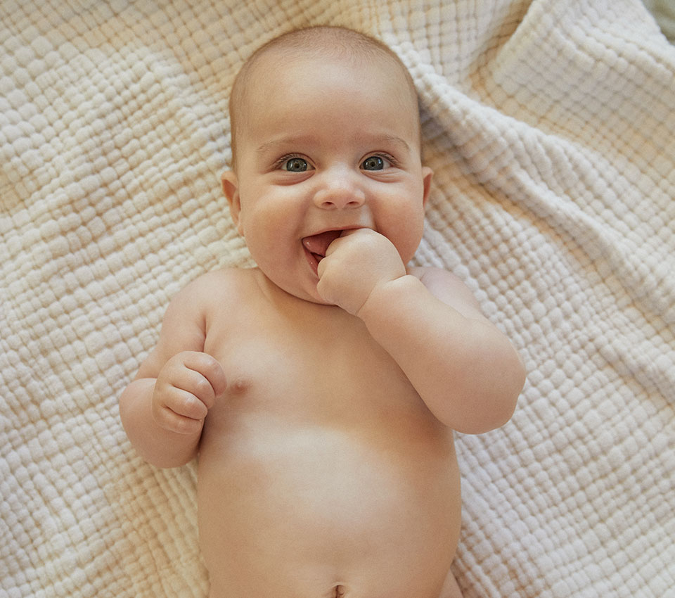 Smiling baby lying on soft blanket with hand in mouth.